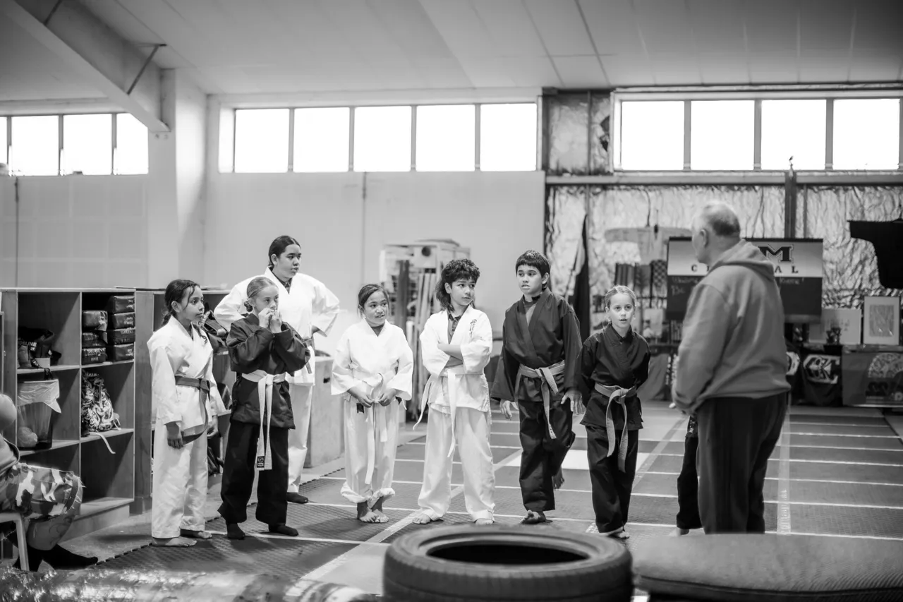 A wide view of seven students lined up across the mats facing their kaiako, under the gym high windows
