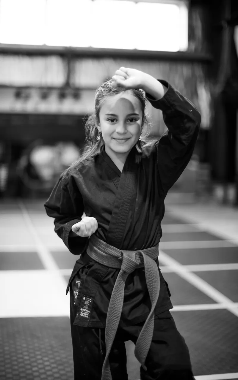A grey-belt student raises one fist above her head in a rising block, the other hand guarding at her waist